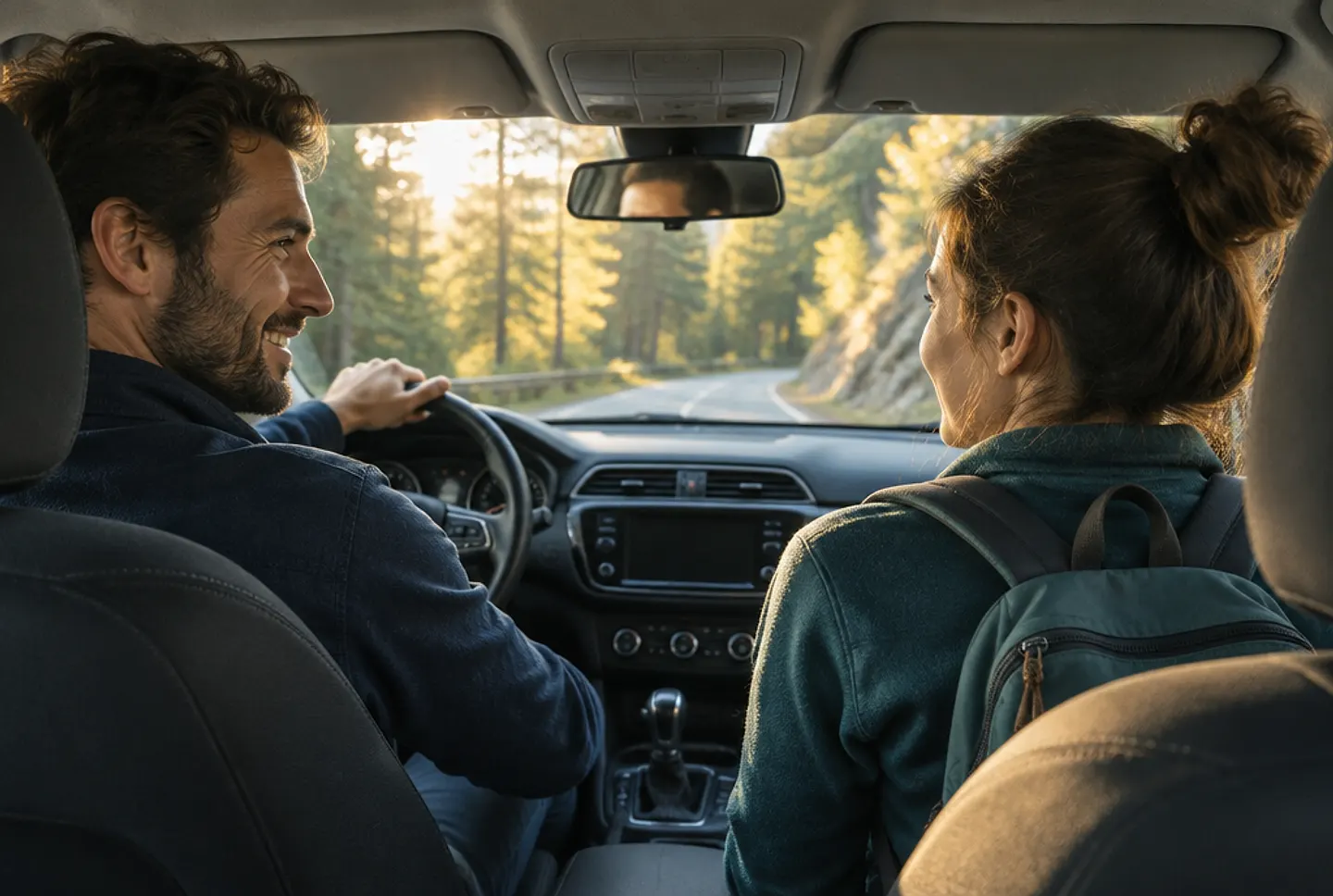 A driver smiling at his passenger as they drive along a sunlit forest road